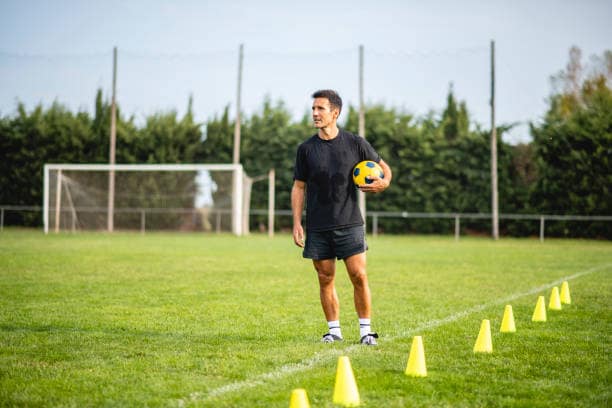 Fit Caucasian male footballer in early 40s standing on field wearing black sports shirt and shorts, holding ball, and looking away from camera.
